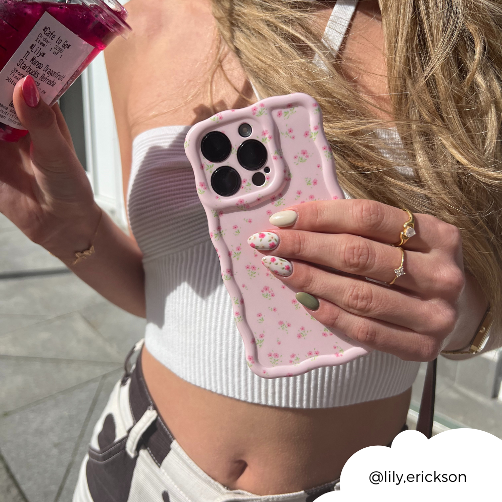 A person with long blonde hair is holding a phone adorned with Coconut Lane's Wavy Phone Case - Ditsy Floral Pink in one hand and a jar of a pink beverage in the other. They are wearing a white crop top and have pastel, floral-patterned nails. Their gold jewelry includes a ring and bracelet. The username