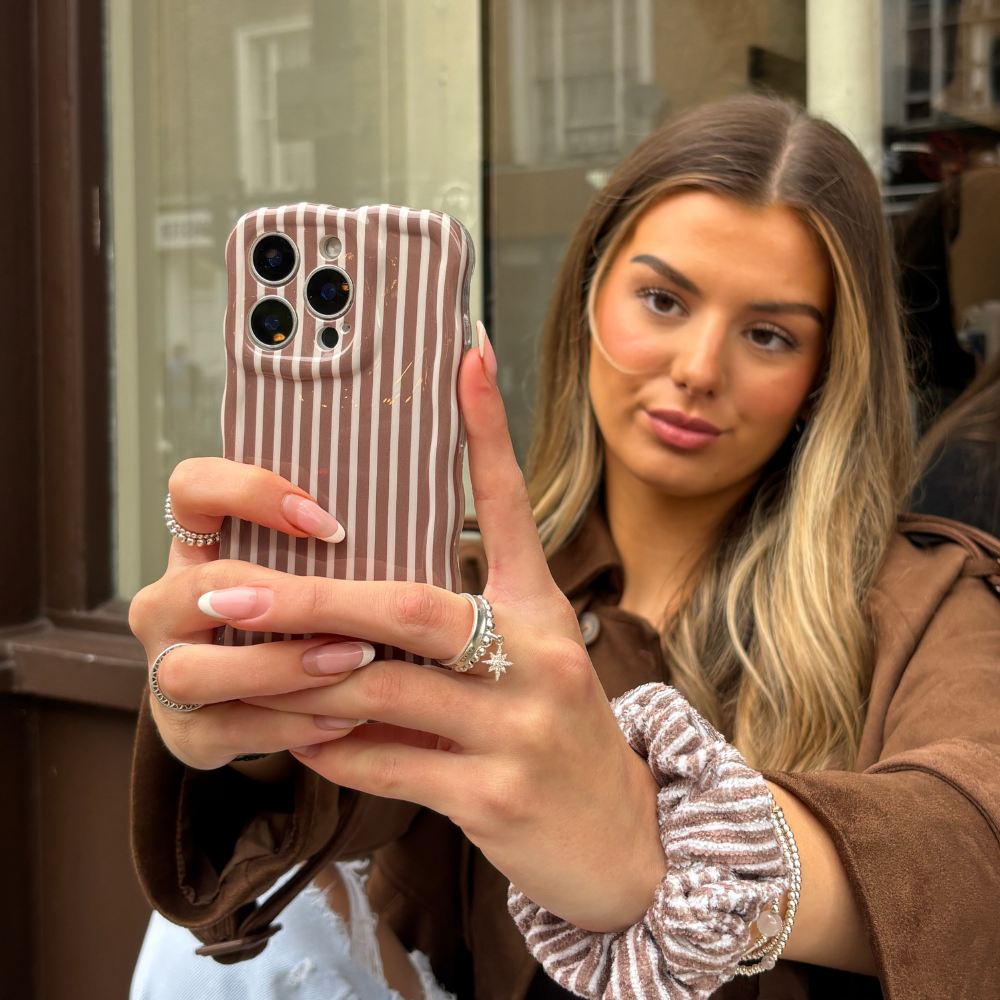 Woman with long hair holds Coconut Lane wavy phone case in chestnut stripes and a matching scrunchie outdoors.