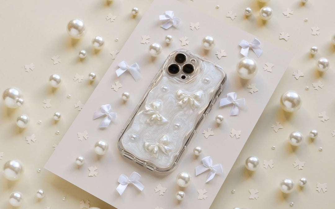 A white iPhone case decorated with small bows and pearls is displayed on a beige background surrounded by scattered pearls, white bows, and star-shaped confetti.