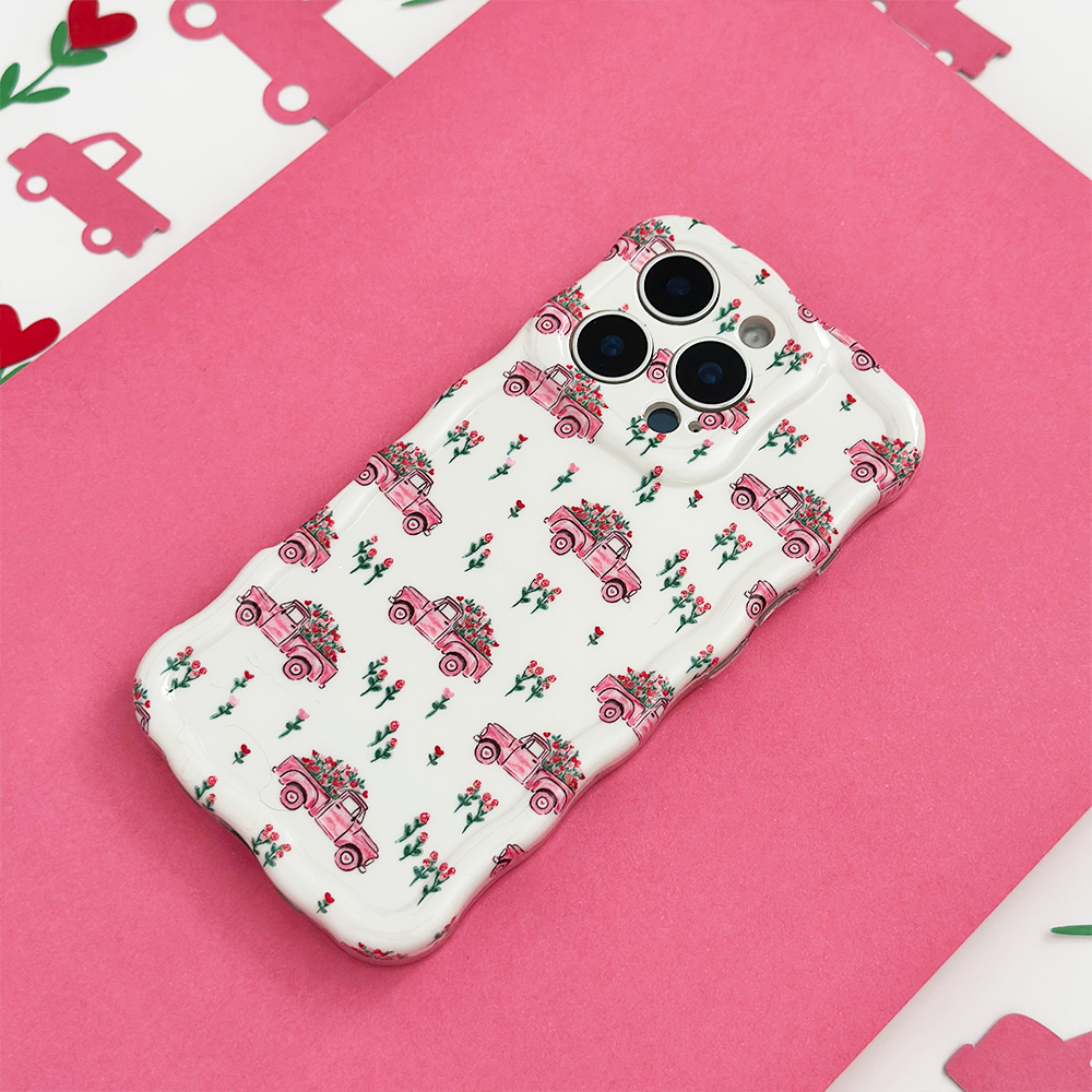 A phone case with a white background and pink vintage car pattern, adorned with small flowers, is placed on a pink surface with matching decorative elements around.