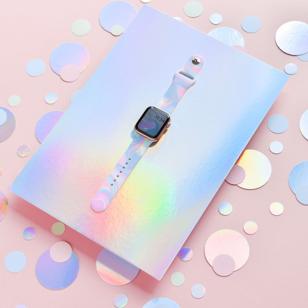 A smartwatch with a colorful, iridescent strap laid on a shiny holographic surface, surrounded by holographic dots. The background is a soft pastel pink, adding a vibrant and playful aesthetic to the scene.