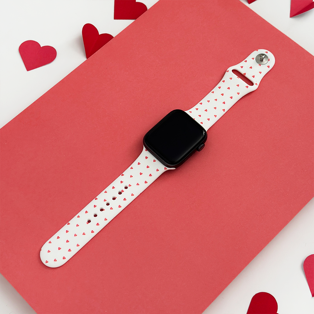 A smartwatch with a white band decorated with small red hearts is placed on a pink surface, surrounded by scattered red and pink paper hearts.