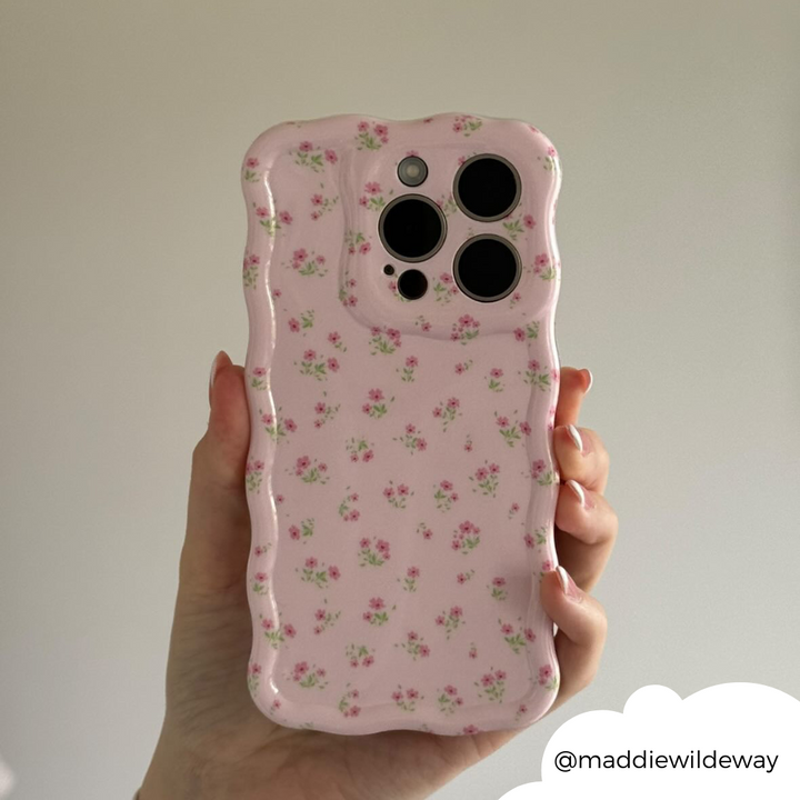 A hand holding a smartphone featuring the Wavy Phone Case - Ditsy Floral Pink from Coconut Lane. The protective pink case is decorated with small, delicate floral patterns and has a distinctive wavy, textured edge that adds a unique touch. The image includes the Instagram handle "@maddiewildeway" in the bottom right corner.