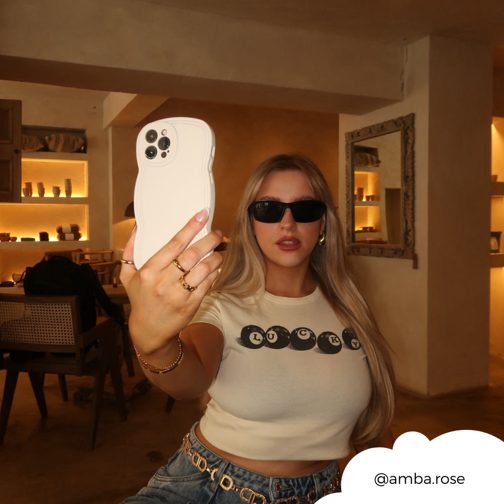 A blonde woman with long hair is taking a selfie with an iPhone encased in a lightweight, highly protective NAKD Curvy Phone Case - Vanilla by Coconut Lane. She is wearing black sunglasses, a white crop top with black text, and blue jeans while standing in a stylish, warmly-lit room. The Instagram handle "@amba.rose" is displayed in the bottom-right corner.