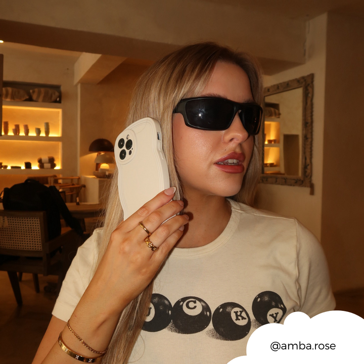 A woman with long blonde hair, wearing dark sunglasses and a white shirt with black graphic letters, holds a huge prop of an iPhone to her ear as if she's talking on it. Behind her is a softly lit room with shelves and a framed mirror. Swipe to shop @amba.rose's favorite NAKD Curvy Phone Case - Vanilla by Coconut Lane—stylish and highly protective!