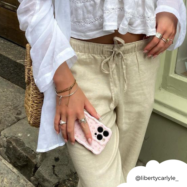 A person wearing beige linen pants and a white long-sleeve top knotted at the waist stands on a stone-paved street. They hold a phone encased in Coconut Lane's Wavy Phone Case - Ditsy Floral Pink, featuring multiple cameras, and have various rings, bracelets, and a straw bag slung over their shoulder. (Credit: @libertycarlyle_).