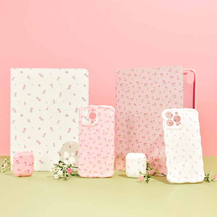 A charming lineup of Coconut Lane tech accessories is showcased against a pastel pink backdrop. The collection features the Ditsy Floral White iPad Case, along with a variety of anti-slip smartphone cases and vegan leather AirPods cases, each embellished with delicate floral designs and accentuated by small white and pink flowers.