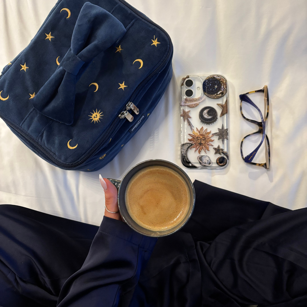 Coconut Lane Clear Phone Case with celestial design lies on the bed near a moon bag, glasses, and a hand holding coffee.