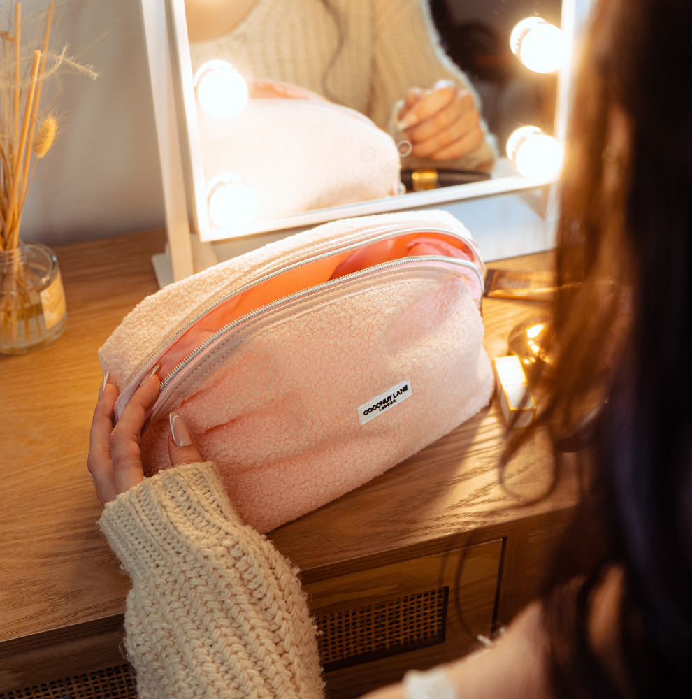 Woman in a cream sweater opens a tan, fluffy Coconut Lane expandable makeup bag at her vanity.