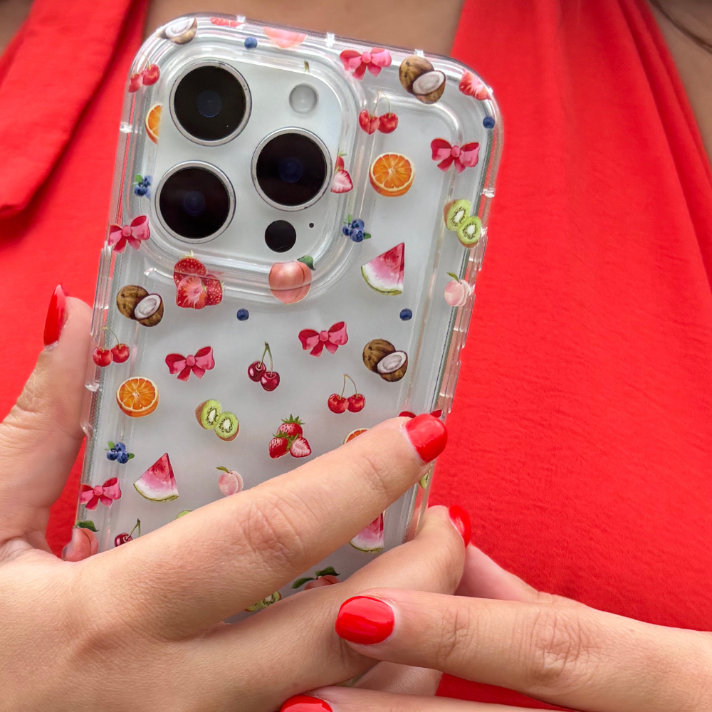 Clear Coconut Lane phone case with colorful fruit design held by a hand with red nails, against a red shirt.
