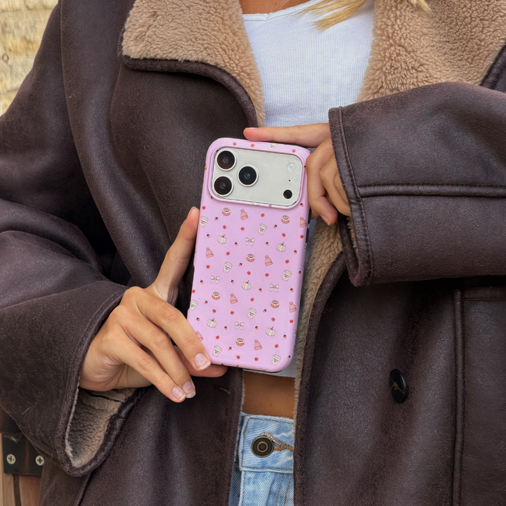 Woman wearing brown aviator jacket holds up an iphone in a baby pink phone case, with small dainty pastries, pumpkins, wooly hats, bows and leaves printed across the case.