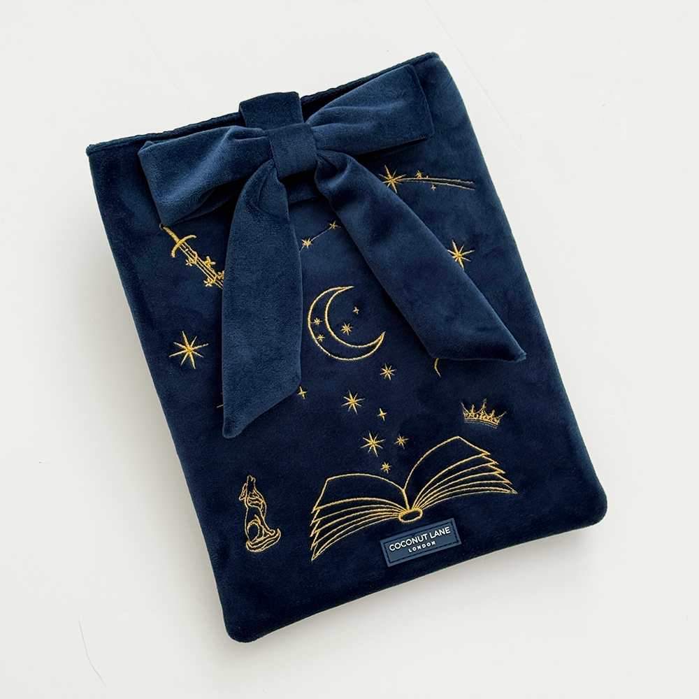 Book Sleeve made from soft navy blue velvet with gold embroidered crescent moon, stars, crown and open book illustrations, finished with a large velvet bow and Coconut Lane logo, photographed on a white background.