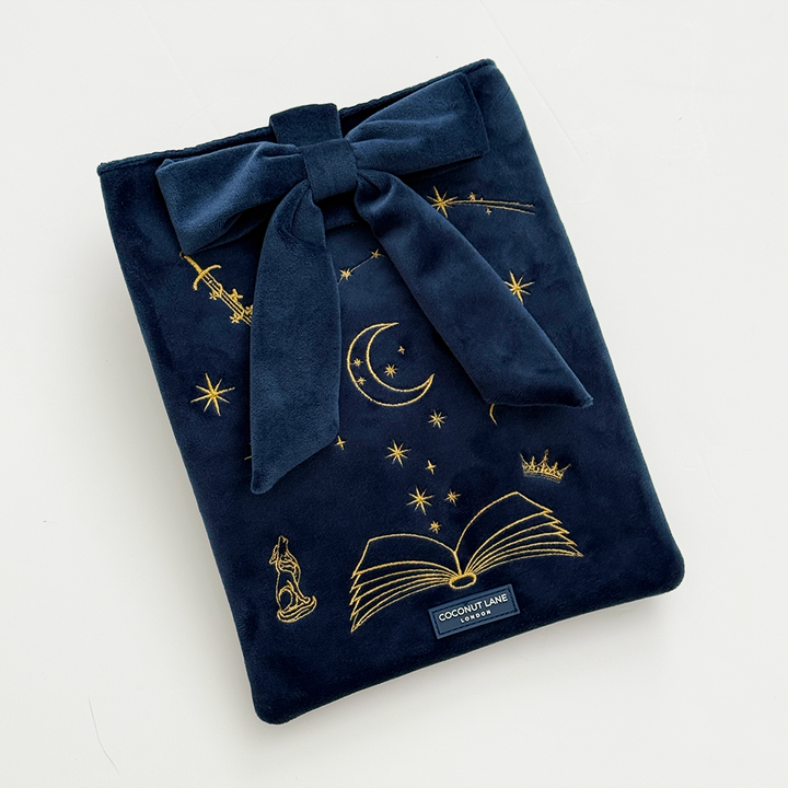 Book Sleeve made from soft navy blue velvet with gold embroidered crescent moon, stars, crown and open book illustrations, finished with a large velvet bow and Coconut Lane logo, photographed on a white background.