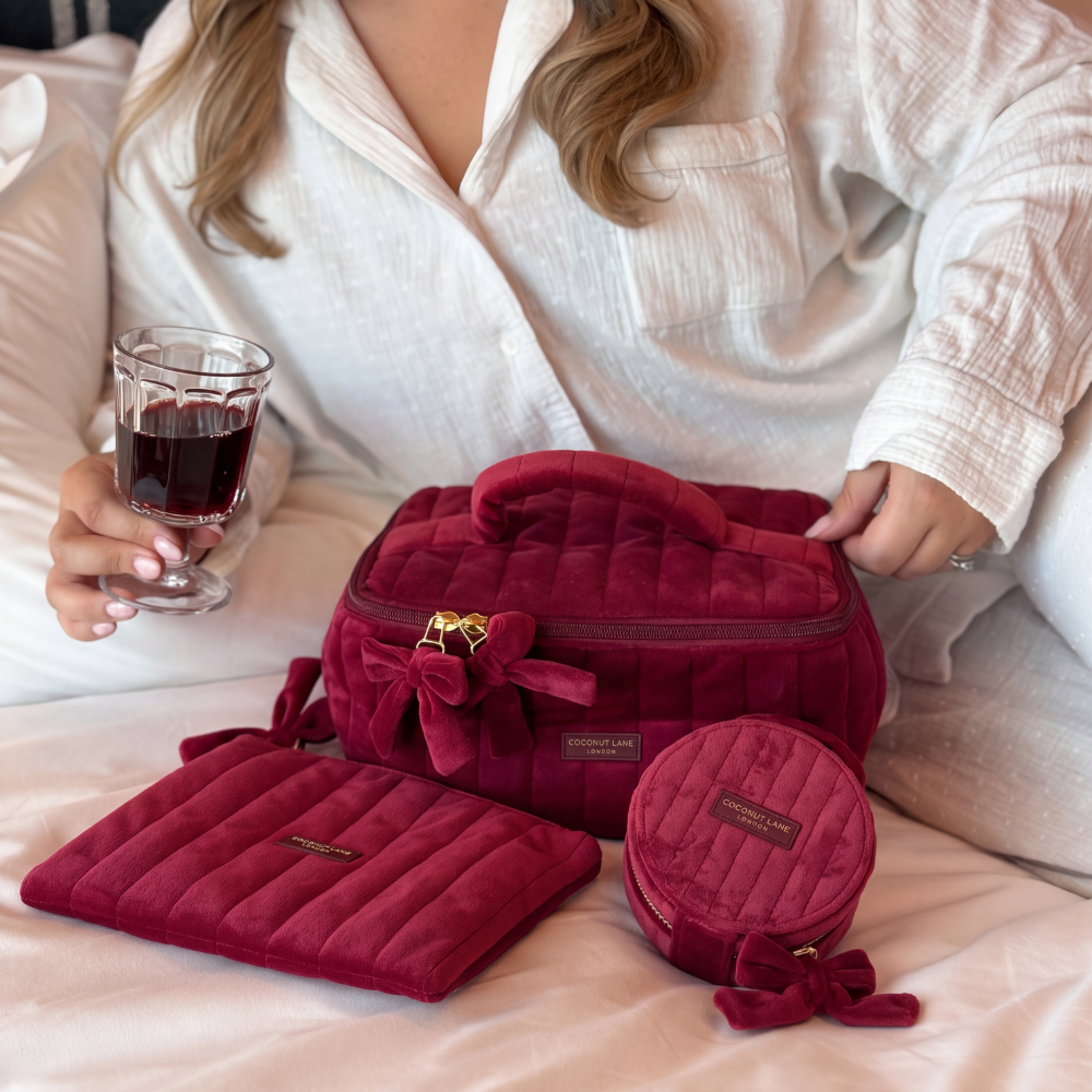 Burgundy velvet jewellery box, vanity case and pouch with a quilted design, top handle, bow detail, and gold-tone hardware. All sit together on a bed with a girl in white Pjs holding red wine