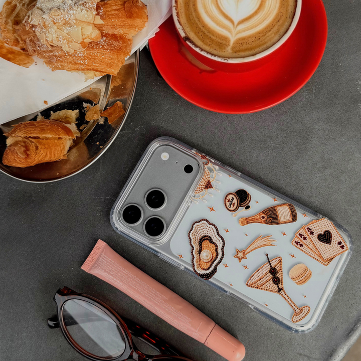 A clear iPhone case with illustrations of a gold-toned cocktail, playing cards, and clam with pearl, shown in a styled flat-lay on a grey surface next to a cup of latte, pastries, and eyeglasses.