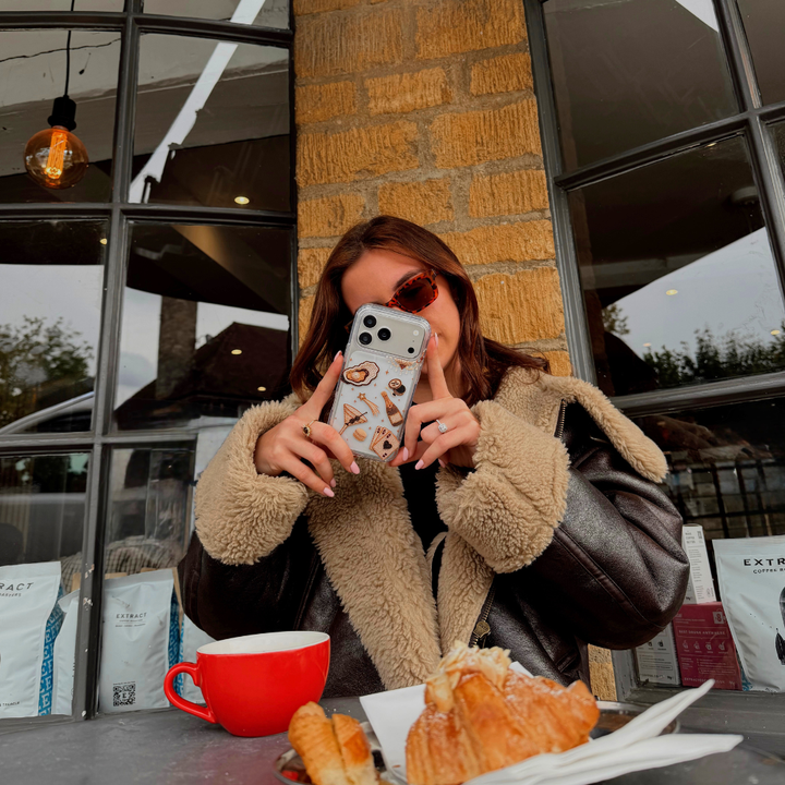 A woman wearing a brown aviator jacket taking a photo with her phone in a cafe. In the foreground, a red coffee mug, pastries, and bags of coffee beans are visible on a table. The phone has a clear phone case with illustrations of a gold-toned cocktail, playing cards, and clam with pearl,