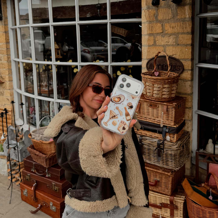 A woman wearing sunglasses and a brown aviator jacket holds up a clear phone case decorated with  illustration of a gold-toned cocktail, playing cards, and clam with pearl. Posing in front of a rustic shop with wicker baskets.