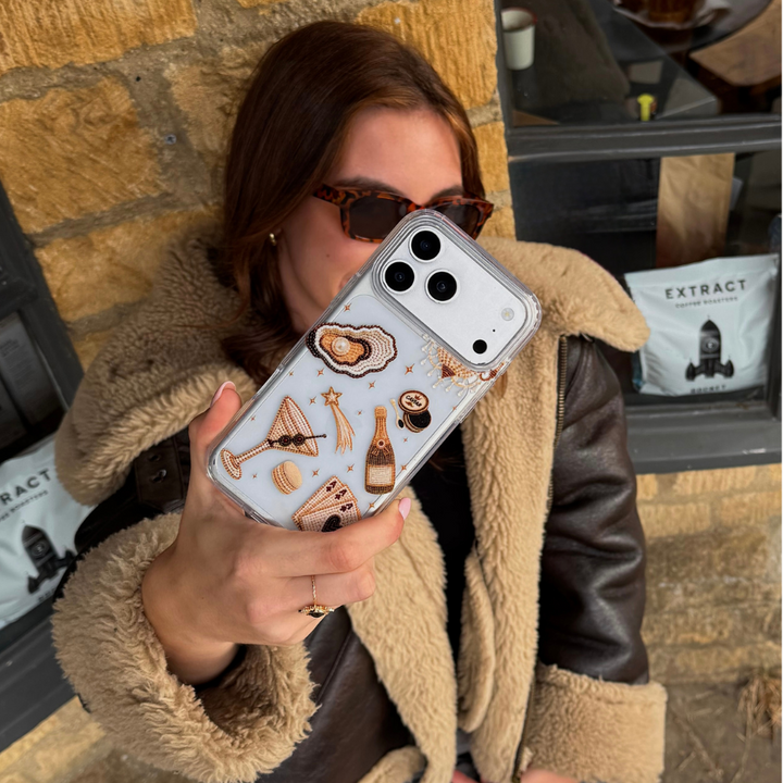 A woman wearing a brown aviator jacket taking a photo with her phone in a cafe. The phone has a clear phone case with illustrations of a gold-toned cocktail, playing cards, and clam with pearl.