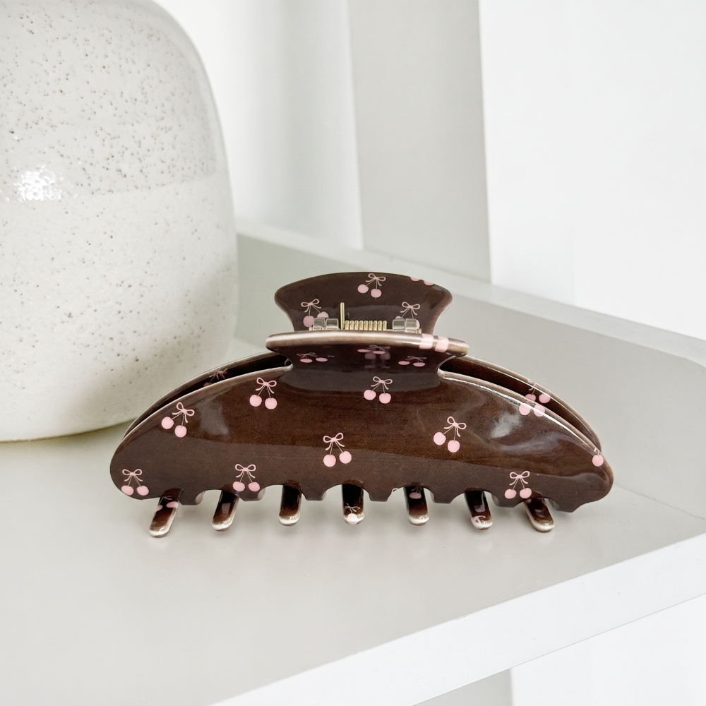 Glossy brown claw hair clip with a pink cherry and bow print, resting on a white shelf.
