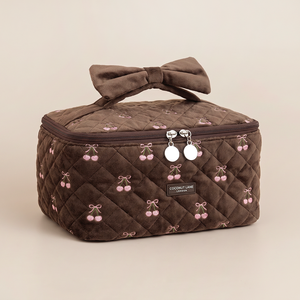 Brown quilted velvet vanity case with pink embroidered cherries, a velvet bow handle and a “Coconut Lane London” label.