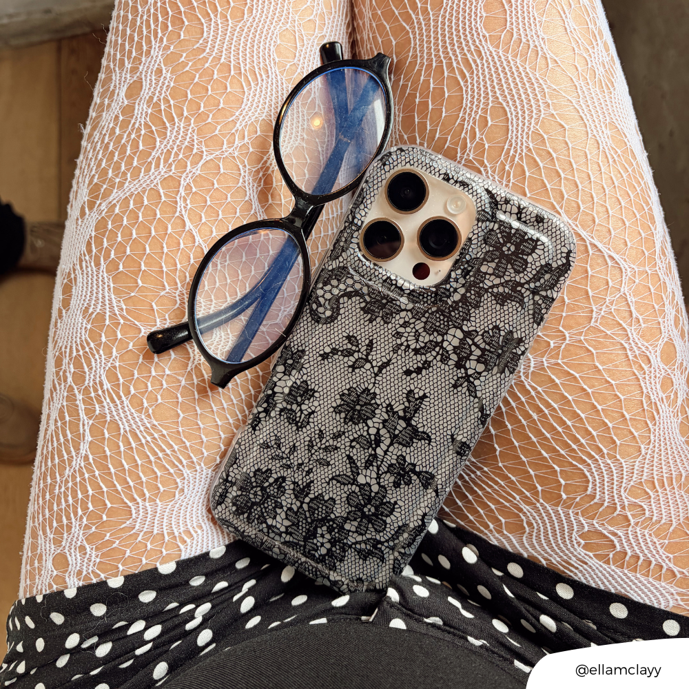 Clear Coconut Lane phone case with black lace design, placed beside eyeglasses on white lace tights and polka dot skirt.
