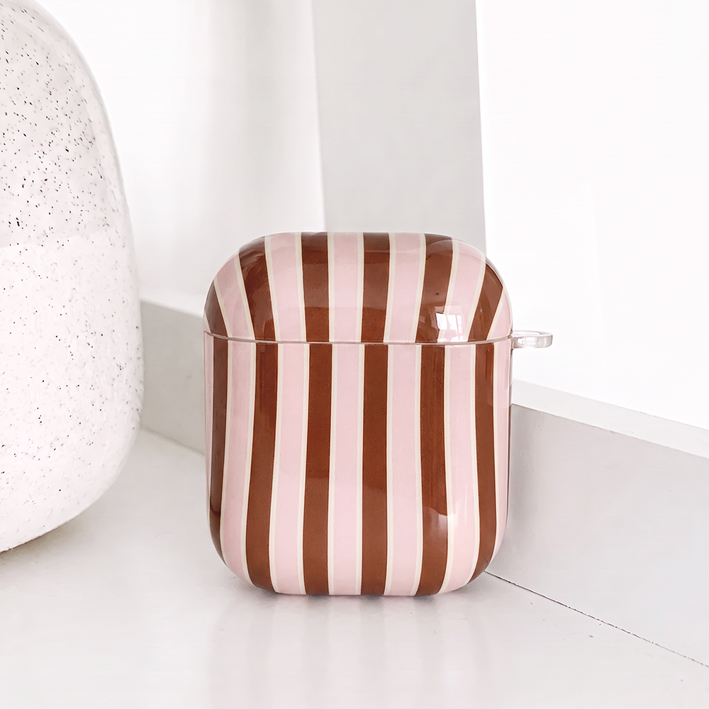 Glossy AirPods Case cover with vertical stripes in pink and brown, separated by thin white lines, with a small clear clip loop on the side. It sits on a white shelf beside a white speckled vase.