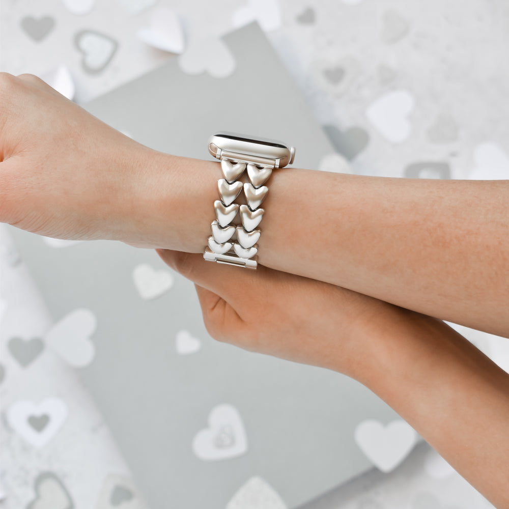 Close-up of a person wearing a Coconut Lane Hearts Chain Apple Watch Strap - Starlight, made of heart-shaped links. The background features a grey surface scattered with various sizes of heart shapes in white and light grey tones, creating a romantic atmosphere. This strap fits ALL Apple Watch models.