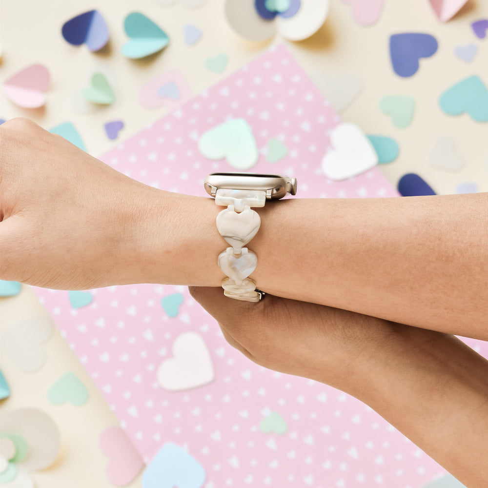 Close-up of a person's wrists wearing an Apple Watch adorned with the Coconut Lane Luxe Hearts Apple Watch Strap - Rose Quartz. The background showcases scattered paper hearts in various vibrant colors and a pink polka-dotted paper, creating a light and whimsical atmosphere. Upgrade your device with the Luxe Hearts Apple Watch Strap for added charm and elegance.