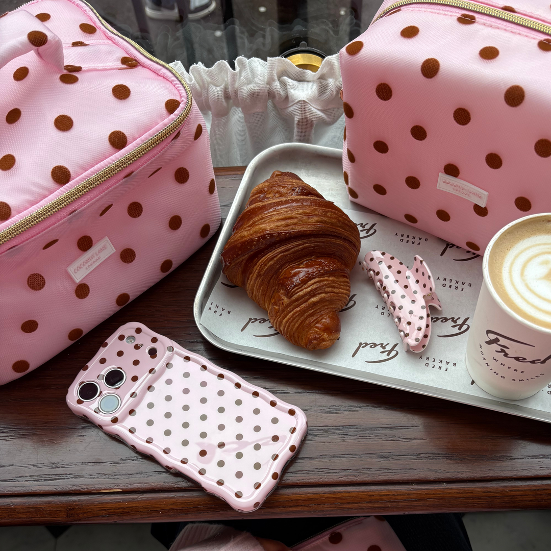 Flat lay photo in a cafe showing the Dot Society pink and brown collection. Including the wavy phone case, pink and brown polka dot claw clip, with a matching makeup bag and vanity case. 