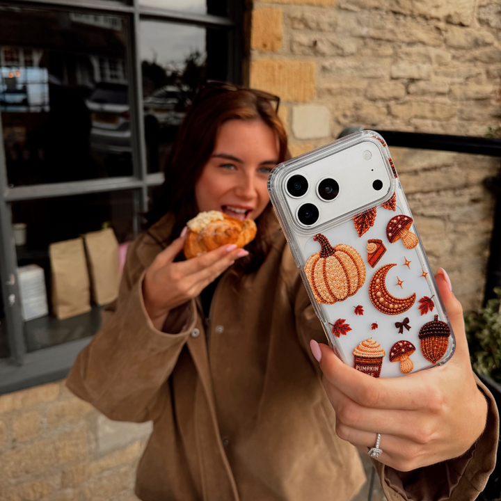 A person holding a clear phone case decorated with an autumn-themed pattern of pumpkins, acorns, leaves, and a pumpkin spice coffee cup, while wearing a brown jacket and smiling in front of a stone building.