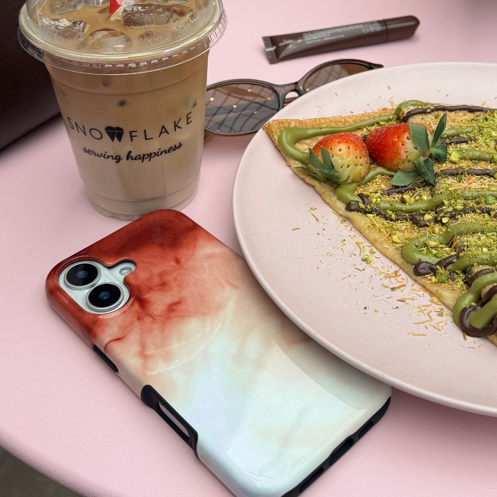 Phone case with a tie-dye design next to a dubai chocolate ctepe and a drink on a pink surface.