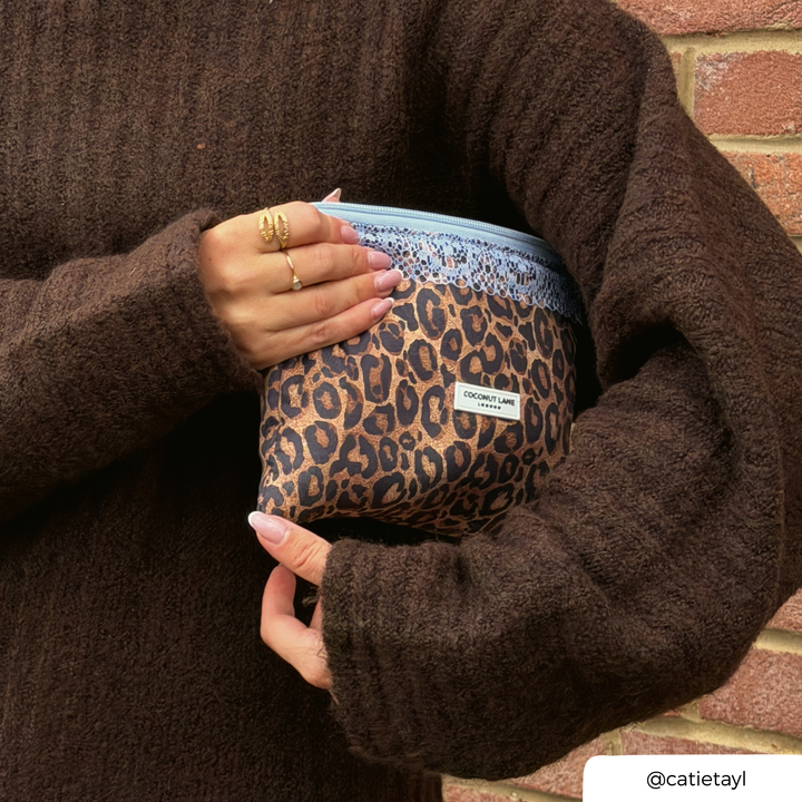 Woman in brown knit jumper, holding the leopard print zip pouch with a pale blue lace trim along the top, a pale blue zip with a silver pull, and a white Coconut Lane London logo badge on the front.