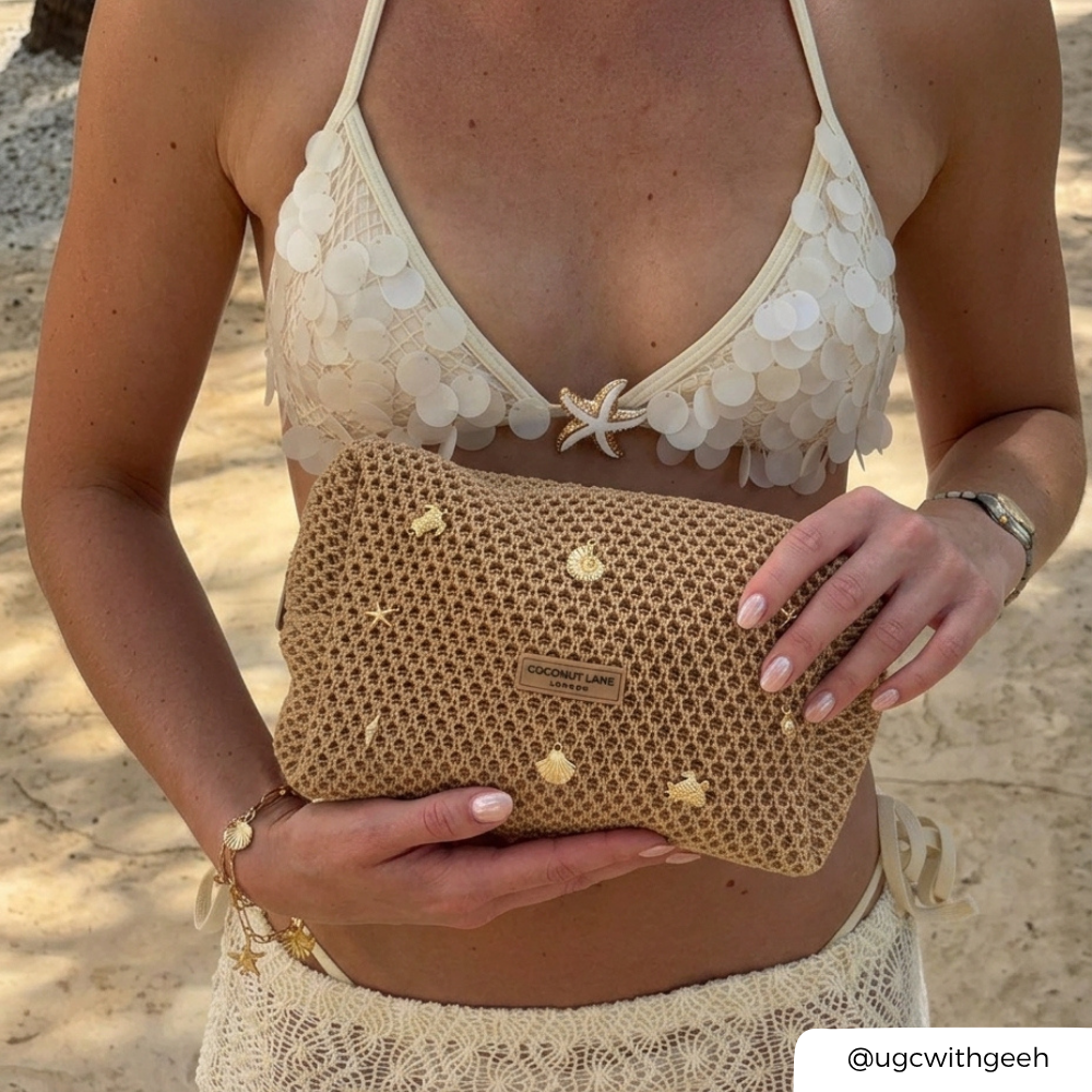 Woman holding a textured beige makeup bag on a sandy background with gold shells