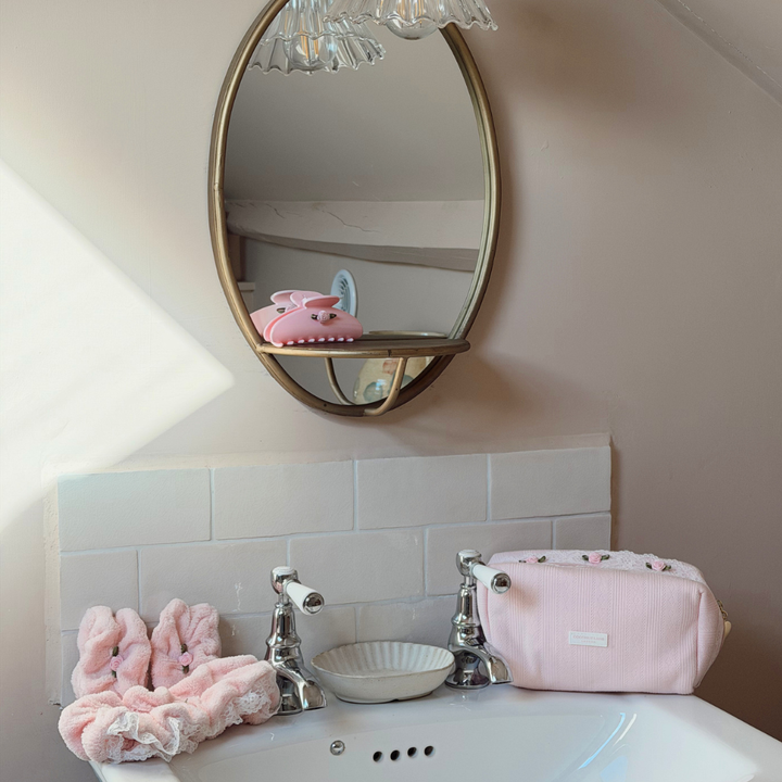 A pink makeup bag with a lace trim and pointelle pattern sits on a white bathroom sink with a matching pink headband and wristband.