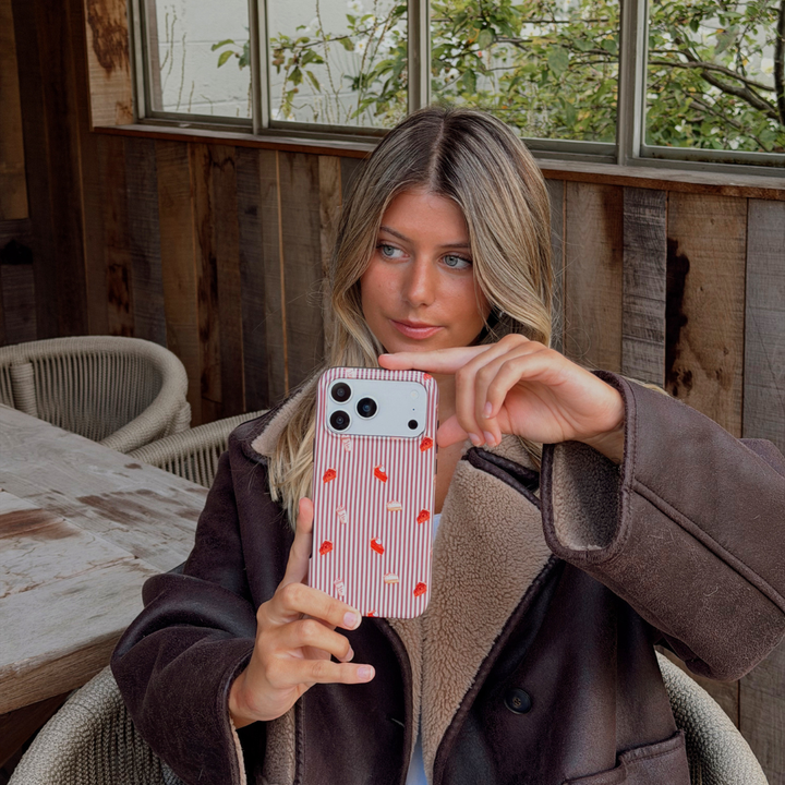 Woman wearing a brown aviator jacket holding up an iphone taking a selfie. The phone is in an autumnal case, with brown vertical stripes, and small prints of pumpkin spice lattes and sweet treats.