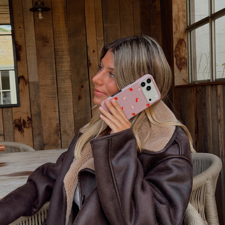 Woman wearing a brown aviator jacket holding up an iphone to her ear. The phone is in an autumnal case, with brown vertical stripes, and small prints of pumpkin spice lattes and sweet treats.