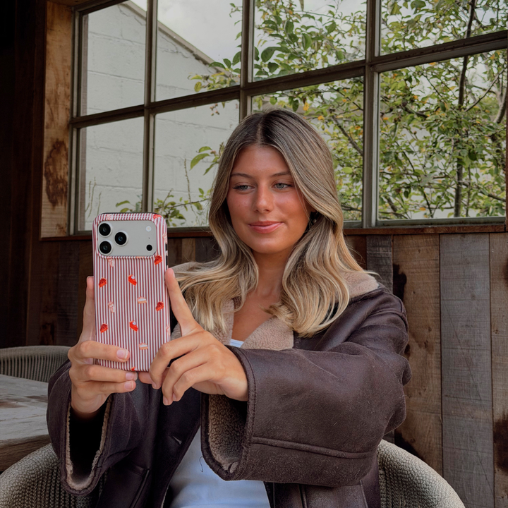 Woman wearing a brown aviator jacket holding up an iphone taking a selfie. The phone is in an autumnal case, with brown vertical stripes, and small prints of pumpkin spice lattes and sweet treats.