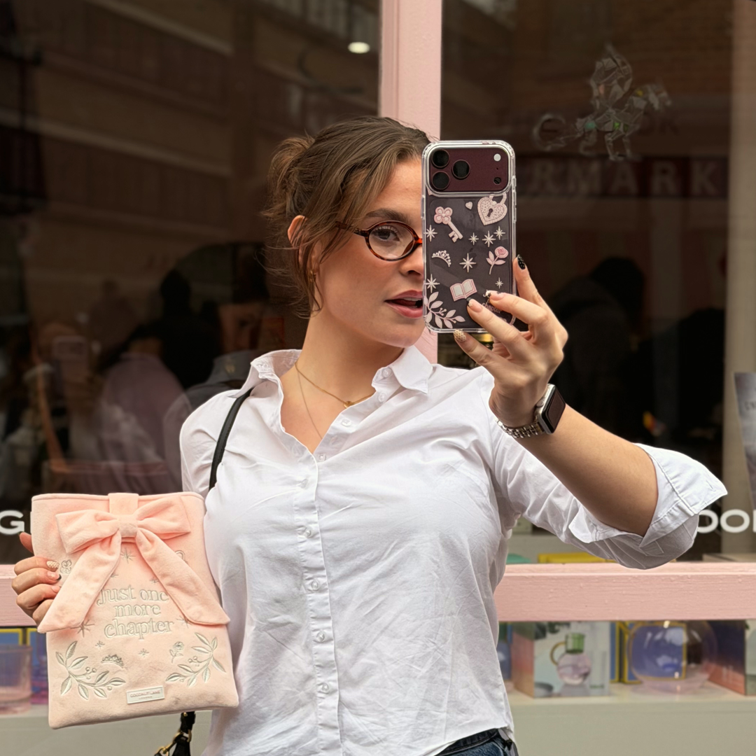 Woman taking a selfie with a burgundy iphone. The iphone is in a Romance Girly Clear Phone Case showing intricate pink key, heart padlock, floral and star details with clear edges.

She is holding a matching Book Sleeve made from soft blush pink velvet with grey embroidered floral details, stars and 'Just One More Chapter' lettering, finished with a large pink velvet bow and Coconut Lane logo, 
