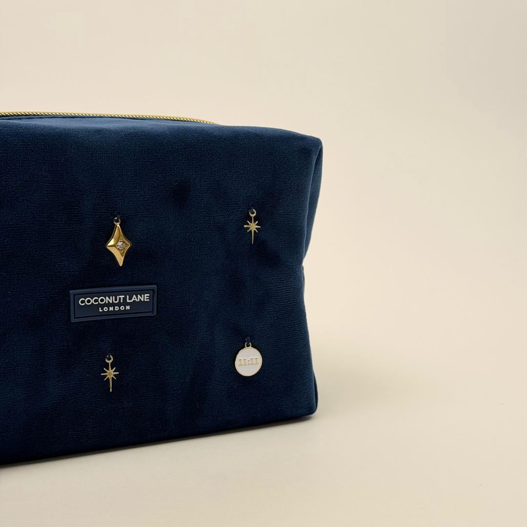 Stardust After Dark makeup bag. Dark navy blue velvet material with gold zip, and sewn-on gold celestial-themed charms including a sun, moon, star, diamond shape and 11:11 charm.  