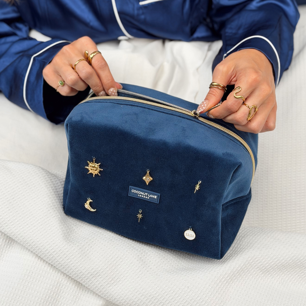 Woman wearing satin navy blue pyjamas holding a Stardust After Dark makeup bag. Dark navy blue velvet material with gold zip, and sewn-on gold celestial-themed charms including a sun, moon, star, diamond shape and 11:11 charm.  