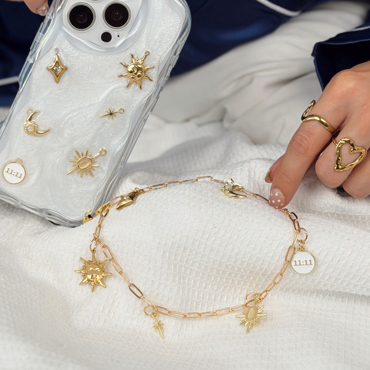 gold colour chain phone strap with celestial style charms, including a moon, sun, star, diamond shape and 11:11 charm. Woman is also holding a matching stardust after dark phone case with matching gold charms.