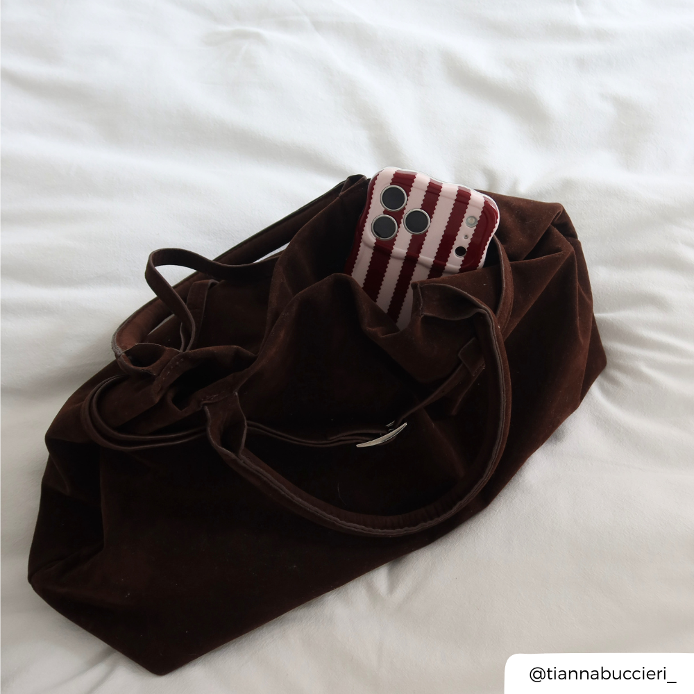 A dark brown velvet tote bag with drawstring straps, shown on white bedding with a phone in a  burgundy and light pink striped wavy phone case peaking out the top of the bag. 