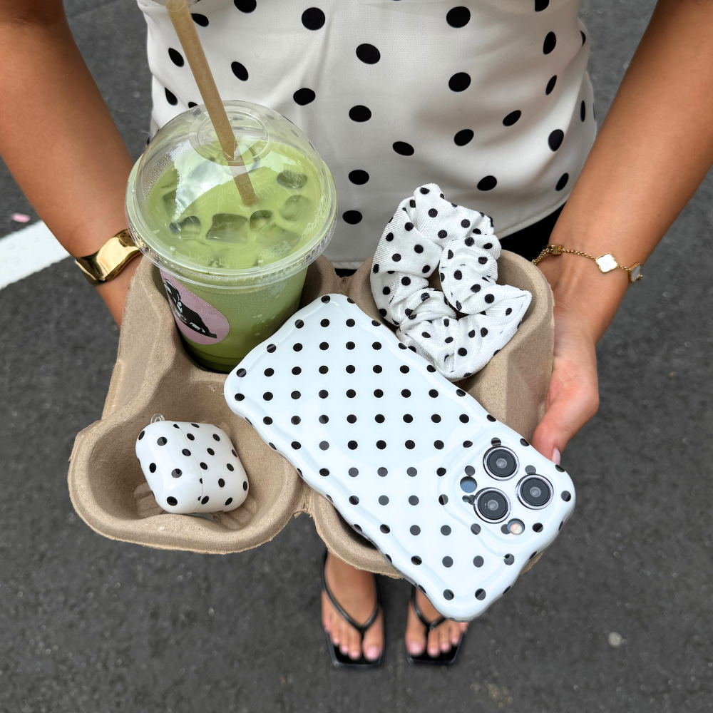 Woman holding tray with green drink, wearing scrunchie, AirPods case, and white polka dot wavy phone case.