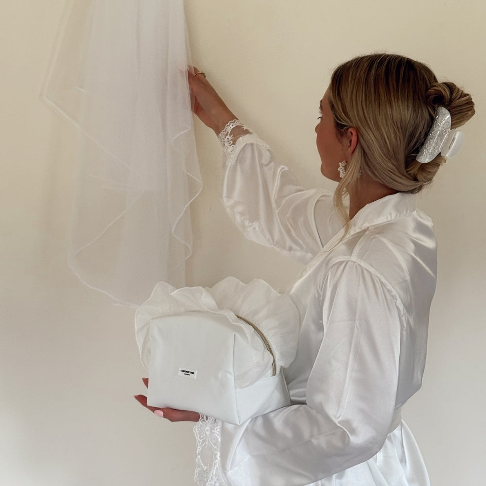 Bride in a white satin robe holding White Ruffle Satin Makeup Bag whilst admiring her veil