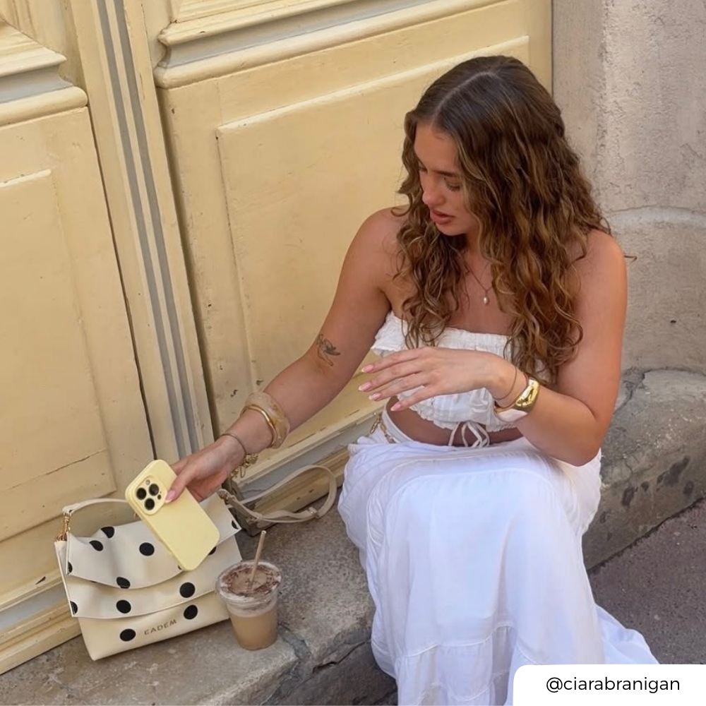 Woman in a white dress sitting on steps with a polka dot handbag and iced coffee.