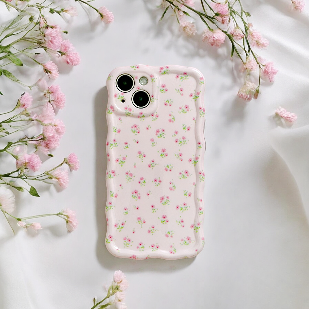 A smartphone with the Wavy Phone Case – Ditsy Floral Pink by Coconut Lane lies on white fabric. Small pink flowers encircle the phone, enhancing the scene with a soft, elegant touch.