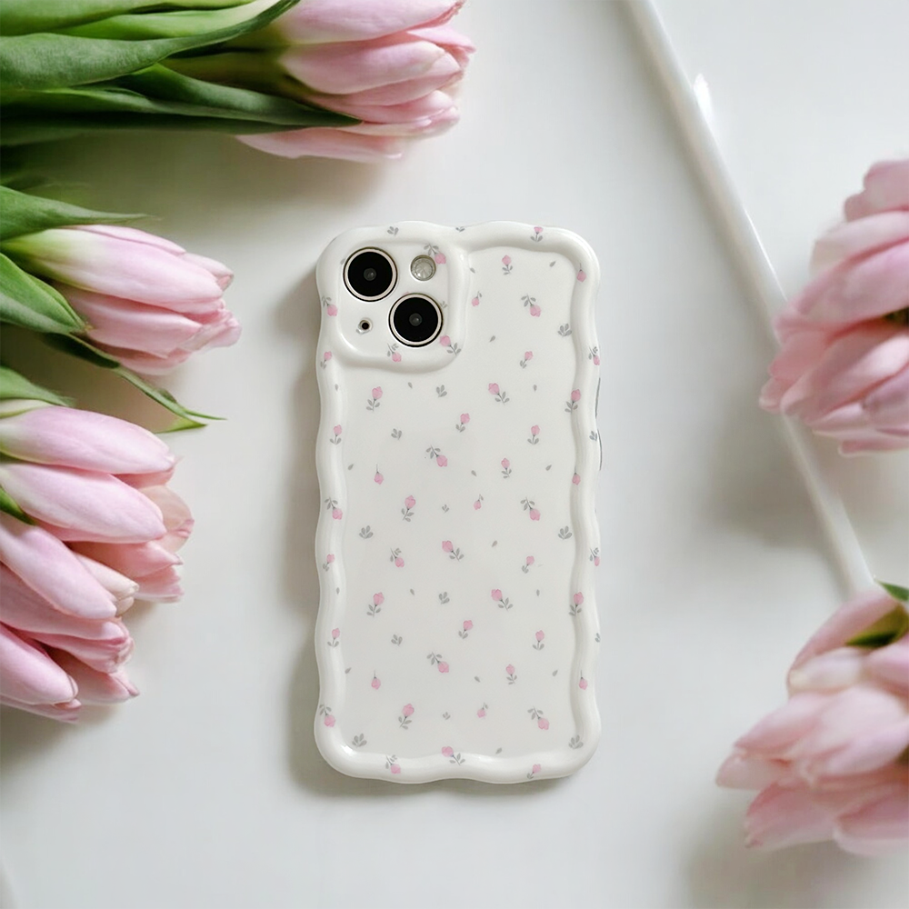 A white smartphone adorned with the Wavy Phone Case - Ditsy Floral White by Coconut Lane, featuring a dainty, scalloped edge and decorated with small pink flowers, is placed on a white surface surrounded by pink tulips. The arrangement exudes a soft and elegant aesthetic.