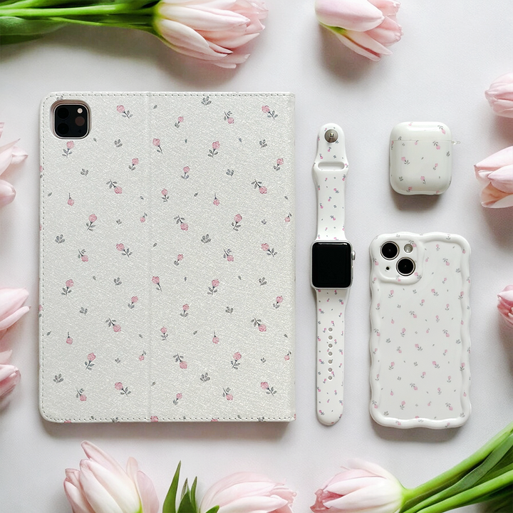 An arrangement showcasing Coconut Lane's Ditsy Floral White iPad Case, a smartwatch, phone case, and wireless earbuds case, all surrounded by delicate pink tulips on a white background. Each tech accessory shares a coordinated white background with a small pink floral design and features anti-slip vegan leather.