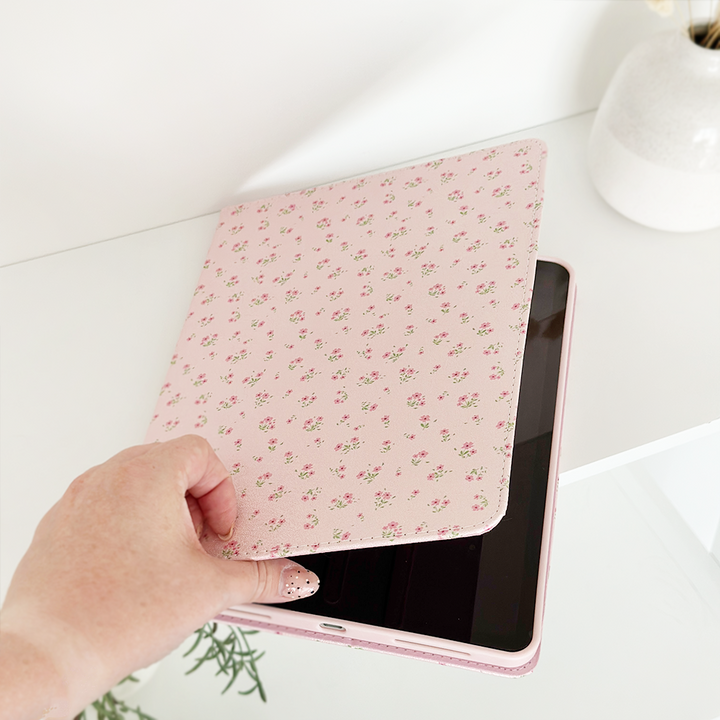 A hand is opening the Coconut Lane Ditsy Floral Pink iPad Case made from vegan leather, revealing a partially visible tablet screen. The scene is bright and clean, with a white vase in the background.