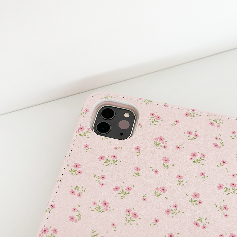 A corner of a smartphone with a Coconut Lane Ditsy Floral Pink iPad Case, showcasing small, delicate flowers in shades of light pink and green, is placed on a white surface. The phone's three rear cameras and flash are visible. This design is also available for the Apple iPad case collection.